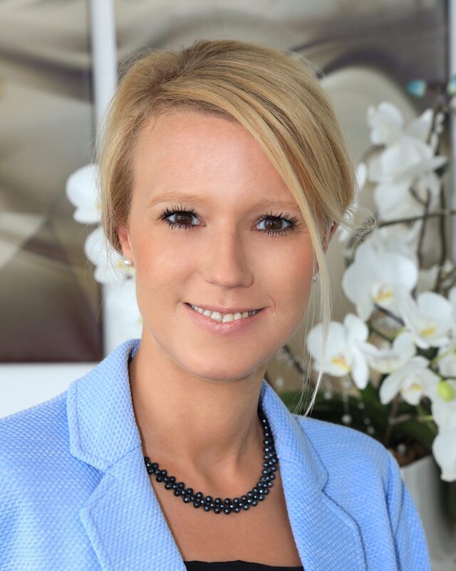 Katrin Hehl, Steuerberaterin: Porträt in hellblauem Blazer mit Perlenkette, dahinter weiße Orchideen